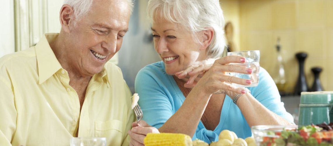 a picture of an older couple smiling at each other at the dinner table because they know what will affect the cost of their dental implant procedures.