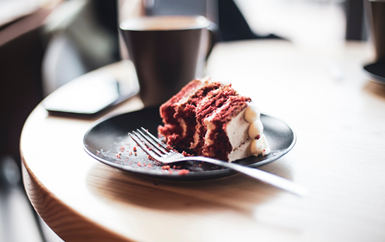 Slice of layered cake on a plate with a fork, placed on a café table next to a cup of coffee