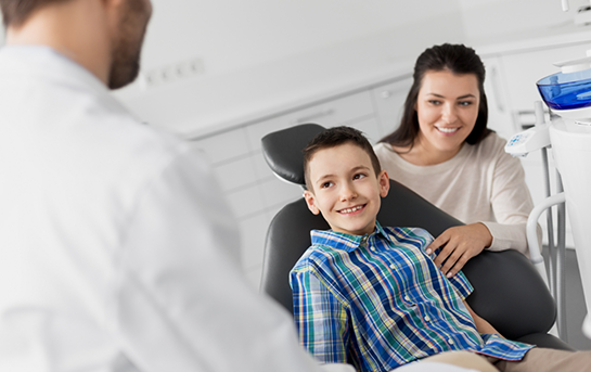 Young boy in the dental chair smiling at his dentist with his mother sitting next to him